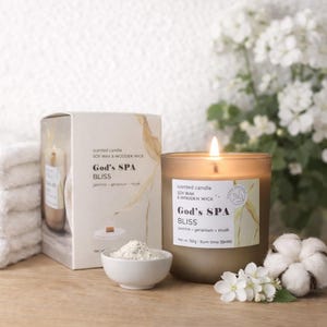 Pode incluir: Uma vela perfumada acesa num frasco de vidro com o texto "God's SPA BLISS" ao lado de uma caixa com o mesmo texto. A vela está ao lado de uma pequena tigela de pó branco, flores brancas e uma bola de algodão.
