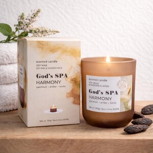 Pode incluir: Uma vela perfumada castanha acesa num frasco de vidro fosco ao lado de uma caixa. A vela e a caixa dizem "God's SPA HARMONY" com "patchouli, âmbar, tonka" como aroma. A caixa e a vela estão numa superfície de madeira.