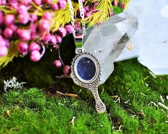 May include: A silver necklace with a small, ornate mirror pendant. The mirror is surrounded by a decorative frame and the pendant hangs from a delicate chain. The necklace is lying on a bed of green moss.