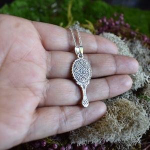 May include: Silver necklace with a detailed ornate mirror pendant. The mirror is round with a decorative handle.