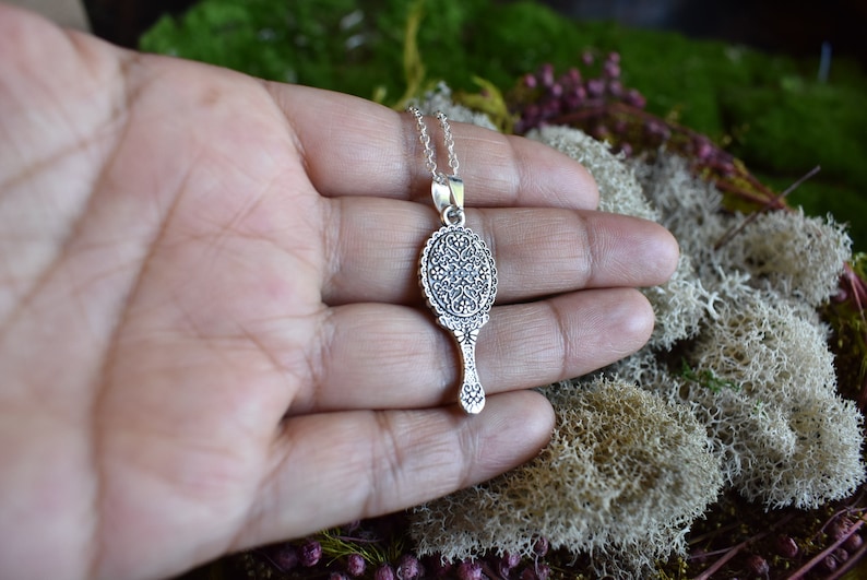 May include: Silver necklace with a detailed ornate mirror pendant. The mirror is round with a decorative handle.
