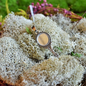 May include: A silver pendant necklace with a round, ornate mirror charm. The charm has a silver frame with a decorative design and a clear glass mirror. The necklace is displayed on a bed of white moss.