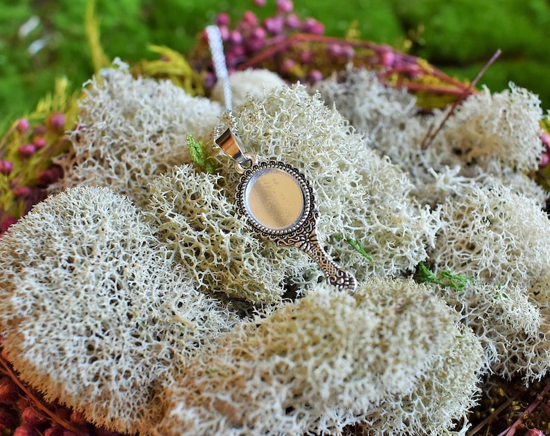 May include: A silver pendant necklace with a round, ornate mirror charm. The charm has a silver frame with a decorative design and a clear glass mirror. The necklace is displayed on a bed of white moss.