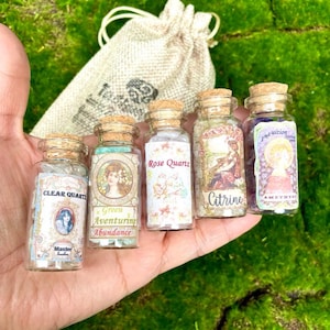 May include: Five small glass bottles with cork stoppers, each labeled with a different crystal name: Clear Quartz, Green Aventurine, Rose Quartz, Citrine, and Amethyst. The bottles are held in a hand, with a small drawstring bag in the background.