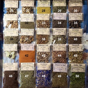 May include: A wooden display case filled with small, labelled packets of dried herbs and botanicals. The packets are arranged in rows and columns, with each labelled with the name of the herb and a number. The colours of the herbs vary, including yellow, brown, and green.
