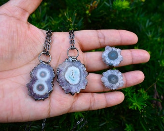 May include: Two necklaces and two pairs of earrings featuring polished, circular gemstones with a purple, white, and blue color scheme. The necklaces have dark metal chains and settings. The earrings are similar in design, without chains.