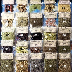 May include: A wooden display case filled with small, clear plastic bags containing various dried herbs and spices. Each bag has a label with the herb's name and weight. The selection includes gardenia fruit, green salt, and lavender.