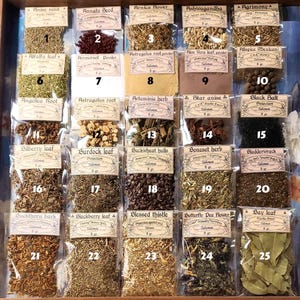 May include: A wooden display case filled with small, labelled packets of dried herbs and spices. The packets are arranged in rows and columns, showcasing a variety of botanical ingredients, including anise seed, arnica flower, and bay leaf.
