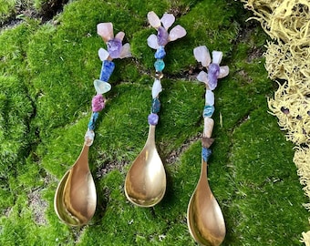 May include: Three gold spoons with colorful gemstone handles. The spoons are arranged on a bed of green moss. The gemstones are various shades of pink, purple, blue, and green.