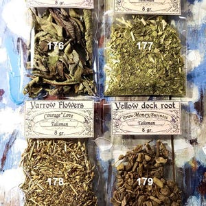 May include: Four small, clear plastic bags filled with dried herbs. Each bag has a label with text such as "Verba Santa leaf" and "Yerba Mate". The labels also include the word "Talisman" and the weight, 8 g. The background is a blue and white painted surface.