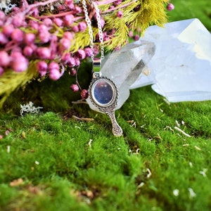 May include: A silver hand mirror pendant necklace with an ornate design. The necklace is displayed on a bed of green moss with pink flowers and a clear crystal.