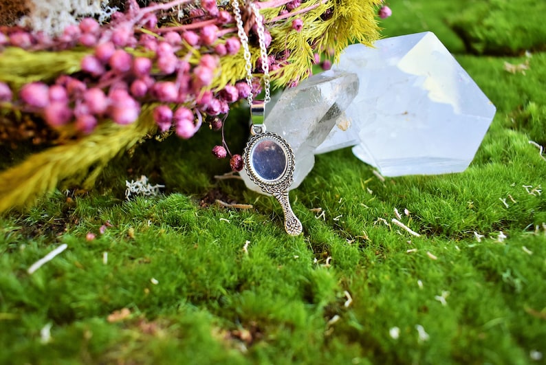 May include: A silver hand mirror pendant necklace with an ornate design. The necklace is displayed on a bed of green moss with pink flowers and a clear crystal.