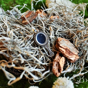 May include: A silver hand mirror necklace with a decorative frame and a black mirror. The necklace is lying on a bed of dried moss and twigs.
