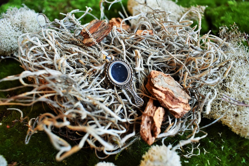 May include: A silver hand mirror necklace with a decorative frame and a black mirror. The necklace is lying on a bed of dried moss and twigs.