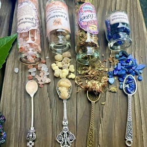 May include: Four small glass bottles filled with various herbs, crystals, and salts, with the labels "Himalayan Pink Salt", "Frankincense", "Red Clover", and "Lapis Lazuli Crystals".  Four ornate spoons with decorative handles are placed in front of the bottles.