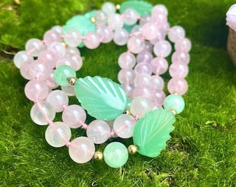 May include: A collection of beaded necklaces featuring pink and green gemstones. The necklaces are strung on pink thread and include leaf-shaped and round beads. The background is a bed of green moss.
