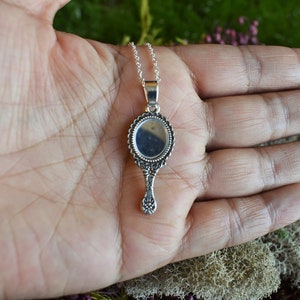 May include: A silver hand mirror necklace with an ornate frame and a round mirror. The mirror is reflecting the hand of the person holding it.