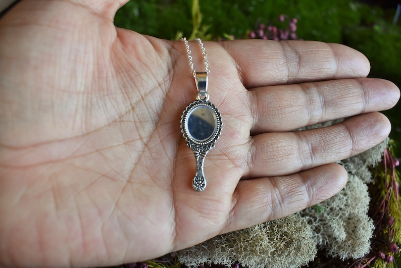 May include: A silver hand mirror necklace with an ornate frame and a round mirror. The mirror is reflecting the hand of the person holding it.