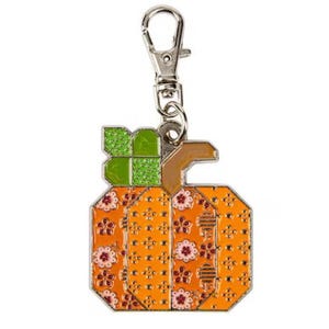 Puede incluir: Un llavero con forma de calabaza con un cierre plateado. La calabaza es naranja con un diseño floral y un tallo verde. Este llavero es de metal y se utiliza para llaves o como accesorio decorativo.