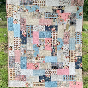 Op de afbeelding: Een patchworkquilt met een verscheidenheid aan stoffen met patronen, waaronder bloemen-, vlinder- en geometrische ontwerpen. De quilt heeft een kleurenpalet van roze, blauw, crème en marine. De quilt hangt buiten aan een waslijn.