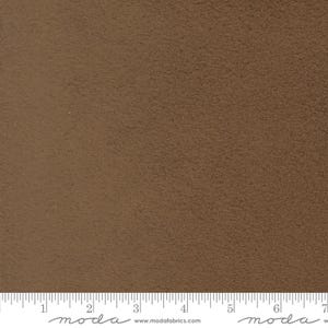 May include: Close-up of a textured, solid brown fabric. The fabric has a slightly rough surface and a warm, earthy tone. A ruler is at the bottom of the image, with the word "moda" and the website address www.modafabrics.com.