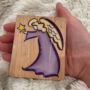 May include: A wooden stamp featuring a cartoon angel in a purple robe with yellow wings and a halo. The angel is holding a yellow star. The stamp is on a light-colored wooden block.