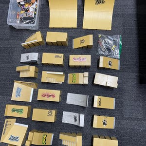 May include: A collection of miniature skateboard ramps and accessories, including two large Tech Deck ramps, various smaller ramps, and a container of tiny skateboards. The ramps are primarily tan and gray, with some featuring graffiti-style designs.