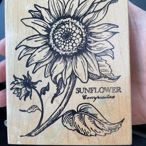 May include: A wooden stamp with a black and white sunflower design. The stamp includes the words "SUNFLOWER" and "Compositae". The rectangular stamp has a natural wood grain and is ideal for crafting or scrapbooking.