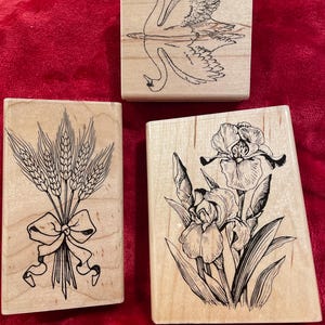 May include: Three wooden stamps with black ink designs. One features a swan reflected in water, another a bundle of wheat tied with a bow, and the third depicts irises. The stamps are on a red background.