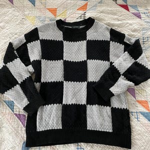 May include: A long-sleeve crew neck sweater featuring a large black and light gray checkered pattern. The knit fabric has a textured, raised dot surface throughout the garment, with solid black ribbed trim at the collar, cuffs, and bottom hem.