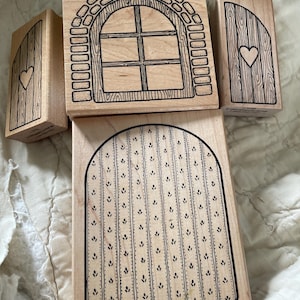 May include: A collection of wooden stamps featuring whimsical door and window designs. The stamps include a window with a brick arch, a door with a heart, and a door with a dotted pattern. The stamps are made of light-colored wood with black ink details.