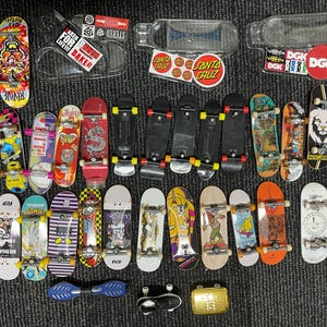 May include: A collection of miniature skateboards in various colors and designs, including graphics and logos. Some boards feature the text "FOR LIFE BAKER", "SANTA CRUZ", and "DGK". Accessories like stickers and a fingerboard are also present.