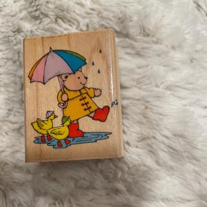May include: A rectangular wooden rubber stamp featuring a colorful illustration of a bear wearing a yellow raincoat and red boots, holding a multi-colored umbrella while walking in a puddle with two small yellow ducks wearing matching red boots.