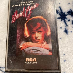 May include: A vintage cassette tape featuring the album "Young Americans" by David Bowie. The cover art showcases a portrait of Bowie with red hair and a cigarette. The RCA logo and catalog number are visible.