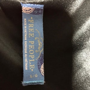 May include: Blue clothing tag with white text that reads "Free People." The tag also says "Made in Vietnam" and "Fabric Made in Vietnam." The tag has a small blue and white design in the top left corner and bottom right corner.