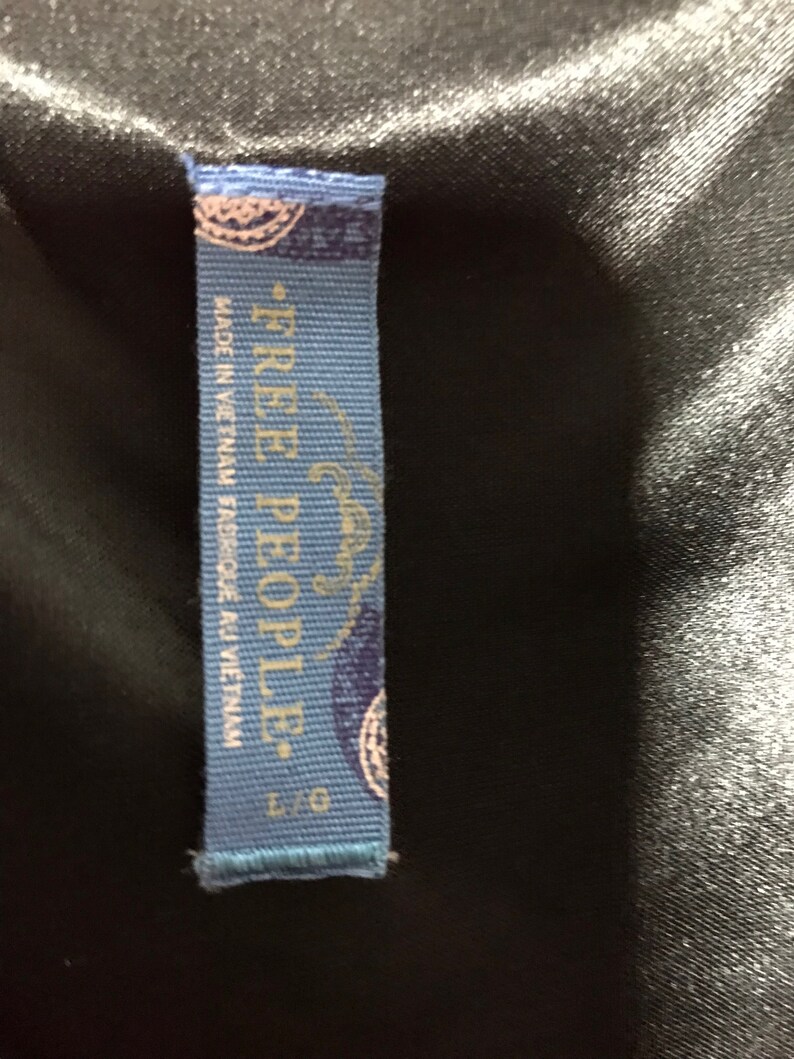 May include: Blue clothing tag with white text that reads "Free People." The tag also says "Made in Vietnam" and "Fabric Made in Vietnam." The tag has a small blue and white design in the top left corner and bottom right corner.