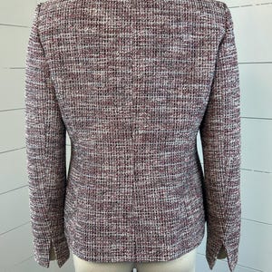 May include: A tweed blazer in burgundy, white, and black. The jacket has long sleeves and a tailored fit. The fabric has a textured appearance. The blazer is displayed on a mannequin, highlighting its classic design and structured silhouette.