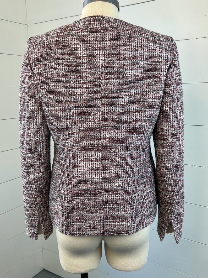 May include: A tweed blazer in burgundy, white, and black. The jacket has long sleeves and a tailored fit. The fabric has a textured appearance. The blazer is displayed on a mannequin, highlighting its classic design and structured silhouette.