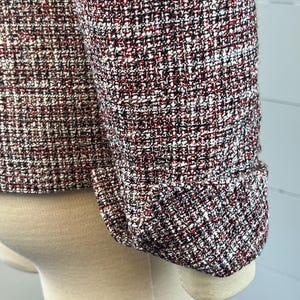 May include: Close-up of a tweed jacket sleeve. The fabric is a mix of red, black, and white threads, creating a textured pattern. The sleeve is cuffed, showing the tailored design of the garment. The jacket is displayed on a mannequin.