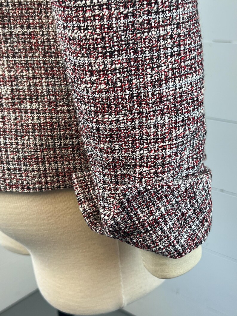 May include: Close-up of a tweed jacket sleeve. The fabric is a mix of red, black, and white threads, creating a textured pattern. The sleeve is cuffed, showing the tailored design of the garment. The jacket is displayed on a mannequin.