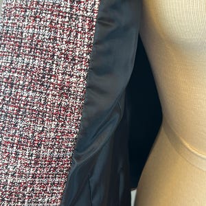 May include: Close-up of a tweed jacket with a black satin lining. The tweed fabric is a mix of red, white, and black threads. The jacket is displayed on a mannequin.