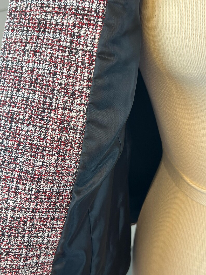 May include: Close-up of a tweed jacket with a black satin lining. The tweed fabric is a mix of red, white, and black threads. The jacket is displayed on a mannequin.