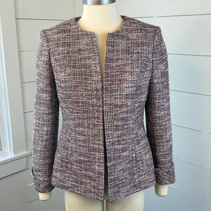 May include: A burgundy, white, and grey tweed jacket with a collarless design. The jacket has long sleeves, a tailored fit, and a button detail on the cuffs. The jacket is displayed on a mannequin.