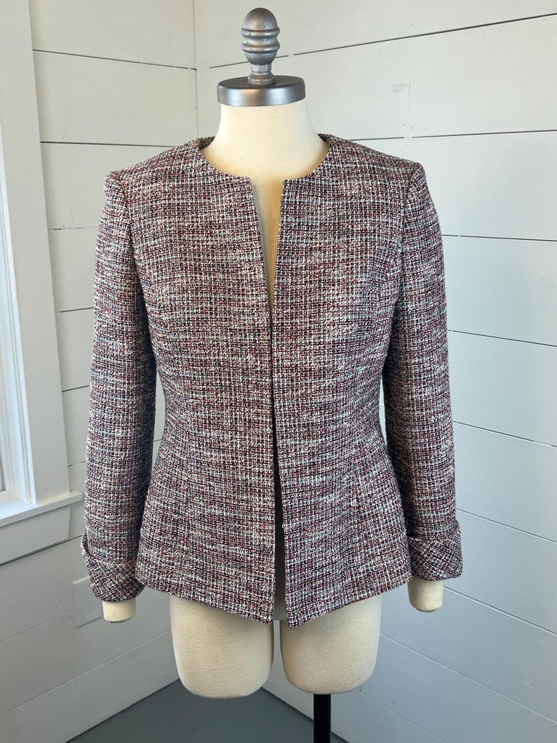 May include: A burgundy, white, and grey tweed jacket with a collarless design. The jacket has long sleeves, a tailored fit, and a button detail on the cuffs. The jacket is displayed on a mannequin.
