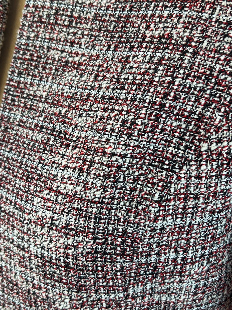 May include: Close-up of a textured fabric with a woven pattern. The fabric features a mix of colours, including red, black, and white, creating a speckled appearance. The weave is tight, suggesting a durable material.