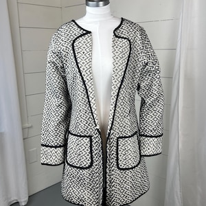 May include: A long, open-front jacket with a black and white geometric pattern. The jacket has black trim along the edges, pockets, and cuffs. It is displayed on a mannequin, showcasing its tailored design and length.