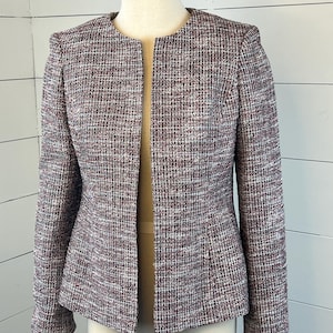 May include: A collarless tweed jacket in a mix of burgundy, white, and grey threads. The jacket has long sleeves and a tailored fit. The garment is displayed on a mannequin, showcasing its woven texture and design.