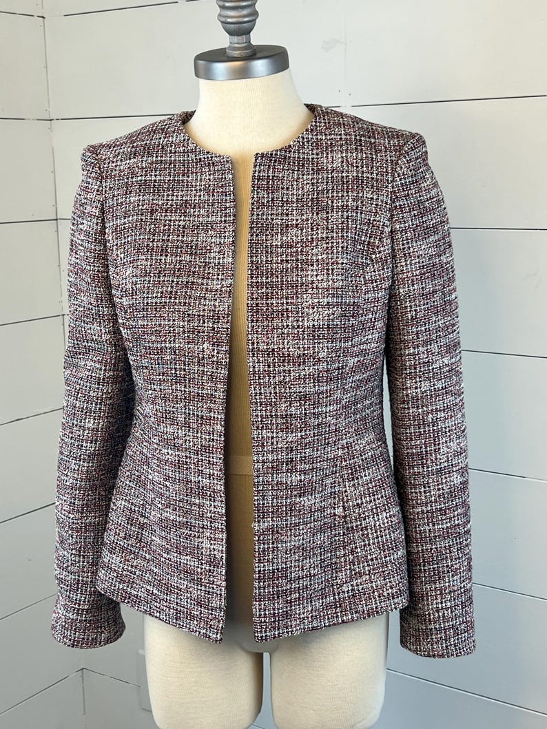 May include: A collarless tweed jacket in a mix of burgundy, white, and grey threads. The jacket has long sleeves and a tailored fit. The garment is displayed on a mannequin, showcasing its woven texture and design.