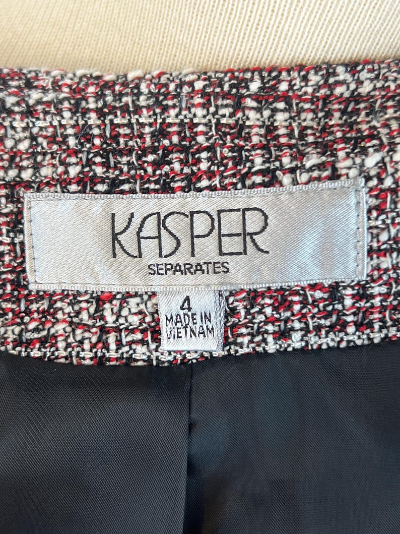 May include: Close-up of a tweed jacket with a black lining. The jacket is a mix of red, black, and white threads. The label reads "KASPER SEPARATES" and "4 MADE IN VIETNAM".