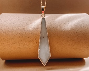 Può includere: Una collana color oro con un lungo ciondolo a punta in pietra bianca traslucida, racchiuso in una sottile cornice di metallo dorato. Il ciondolo pende da una delicata catenina dorata, creando una silhouette geometrica slanciata.